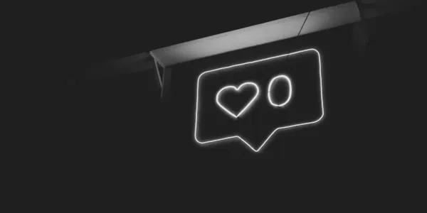 A neon sign shows a speech bubble with a heart and the number zero beside it. This resembles a social media like button. The picture is desaturated and appears in black and white.
