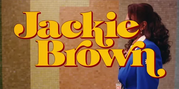 The title card of the movie Jackie Brown. The words 'Jackie Brown' are set in a 1970s stylized font. The fill color of the text is yellow with a thick brown drop shadow emphasizing the character shapes. In the background is an airport scene with the central character. Jackie standing on a travelator (moving walkway) with a mosiac tile wall passing behind her.