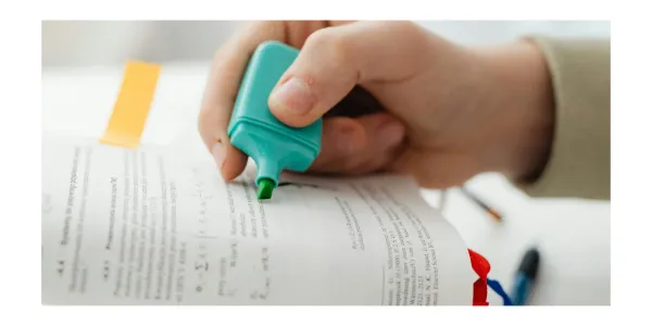 A person has an open book and is highlighting a passage of text with a marker. The page has a sticky post-it note for a reference.