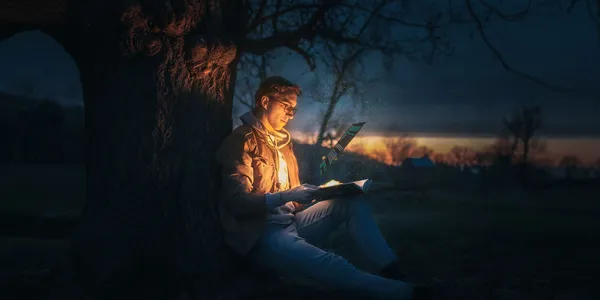 A man is sitting against a tree reading a book at a night. There is an unnatural bright light emanating from the book.