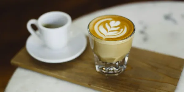 A narrow wooden tray has 2 coffees on it. The first is a small porcelain white cup on a saucer. The second is a small glass containing a latte.