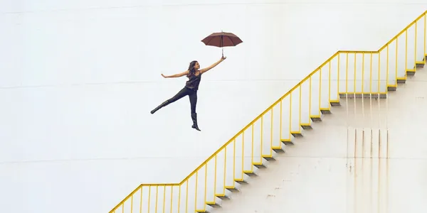 A women in an outstretched pose is holding an umbrella and floating above a set of stairs.